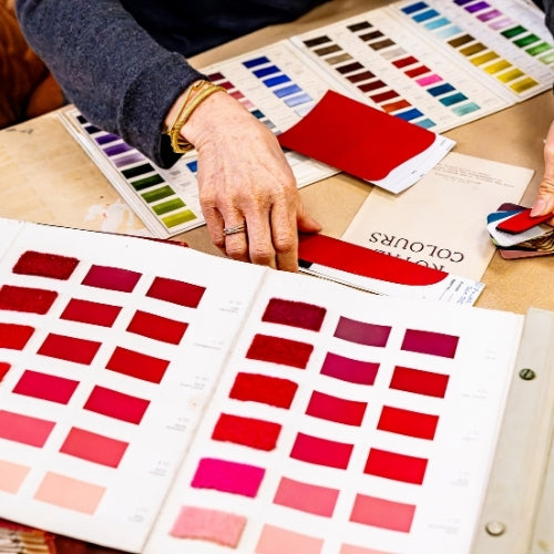 A close-up of a person's hands selecting and comparing fabric color swatches from a large color chart book on a wooden table.