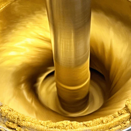 A close-up of a thick, golden yellow paint being stirred in a spiral motion by a vertical metal rod, with sprinkled pigment around the container.