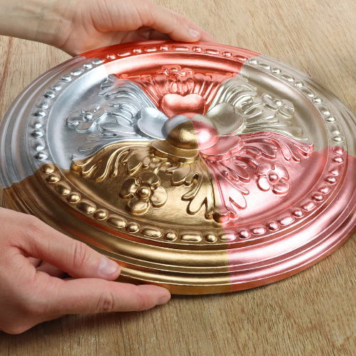 A decorative oval ceiling medallion with embossed floral and scrollwork details, shown being held by two hands on a light wood surface. The medallion has a color-changing finish transitioning from silver to gold to rose gold.