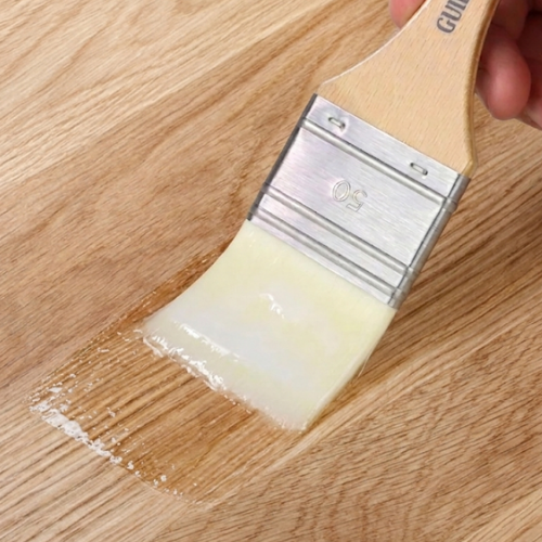 A close-up of a paint brush applying a glossy, pale yellow finish to a wood surface. A hand is holding the wooden handle, which has the brand name 'GULI' partially visible.