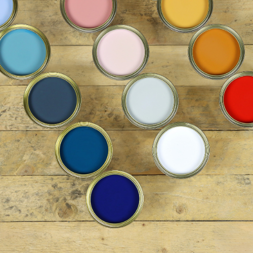 An overhead flat-lay arrangement of open paint cans showing various paint colors, including blue, pink, yellow, orange, red, white, and navy, set on a rustic wooden plank background.