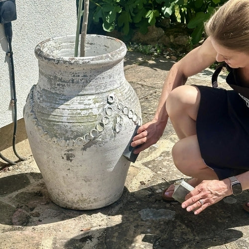 A woman crouches beside a large weathered ceramic pot outdoors, using a sanding block to smooth the rough, distressed surface of the pot.