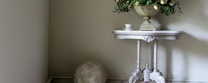 A grey ornate table in a styled interior corner, holding a large stone planter with greenery and a small white cup and saucer. A round stone sculpture sits on the floor beside the table against a beige wall.