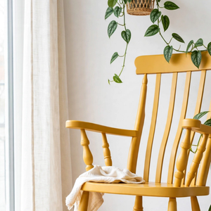 
                      
                        A wooden rocking chair painted in Jubilee Oxford Ochre warm yellow paint, with a linen curtain behind and a trailing plant hanging over it
                      
                    