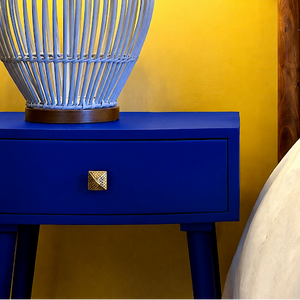 
                      
                        A close-up of a vibrant royal blue bedside table with a single drawer and a textured gold pull handle, topped with a light blue woven basket, set against a bright yellow wall.
                      
                    