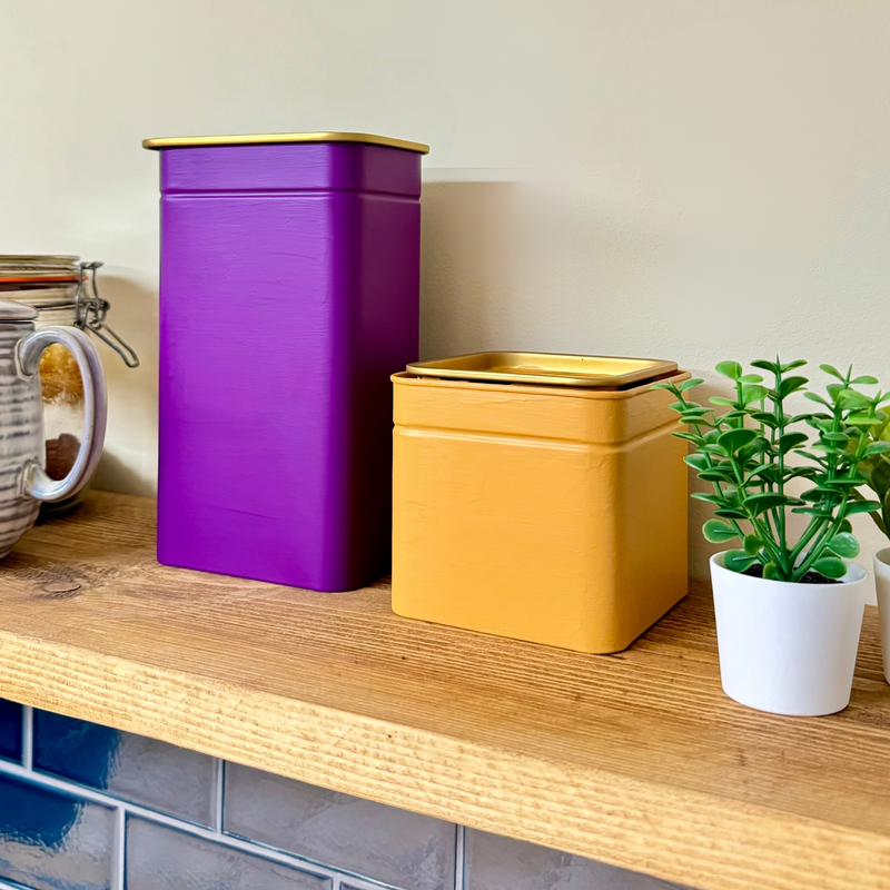 Purple and gold storage canisters on a wooden shelf with a plant and jar.