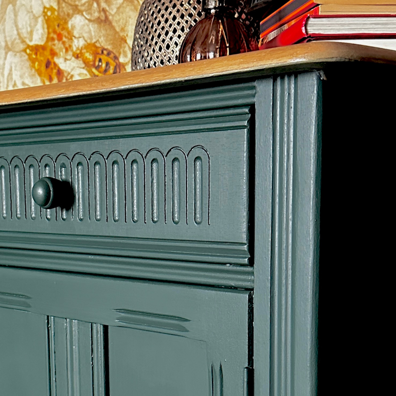 A close-up of a painted wooden cabinet or sideboard in a muted blue-green tone, showing part of a drawer with a round knob, decorative paneling, a wood top, and books and decor on top.