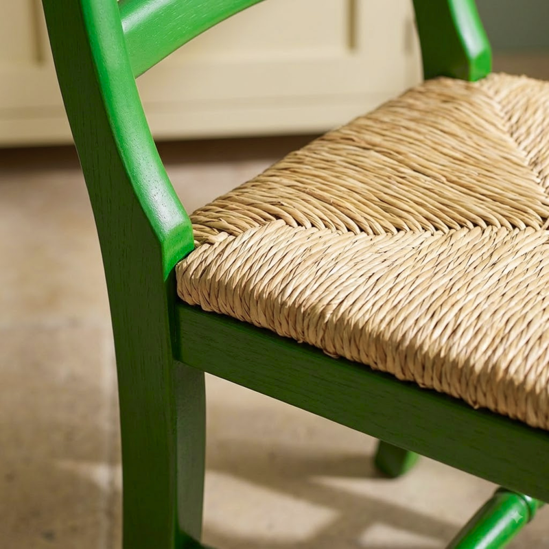 Close-up of a green wooden chair with a woven natural straw seat, shown in a real indoor setting with a blurred floor and wall in the background.