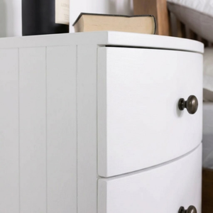 
                      
                        Close-up of a white wooden nightstand or dresser with two drawers and round dark metal knobs, shown in a bedroom setting with a book on top.
                      
                    