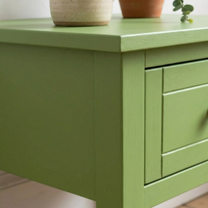 
                      
                        A close-up of a sage green wooden side table or cabinet with a single visible drawer front, shown in a real interior setting with potted plants on top.
                      
                    
