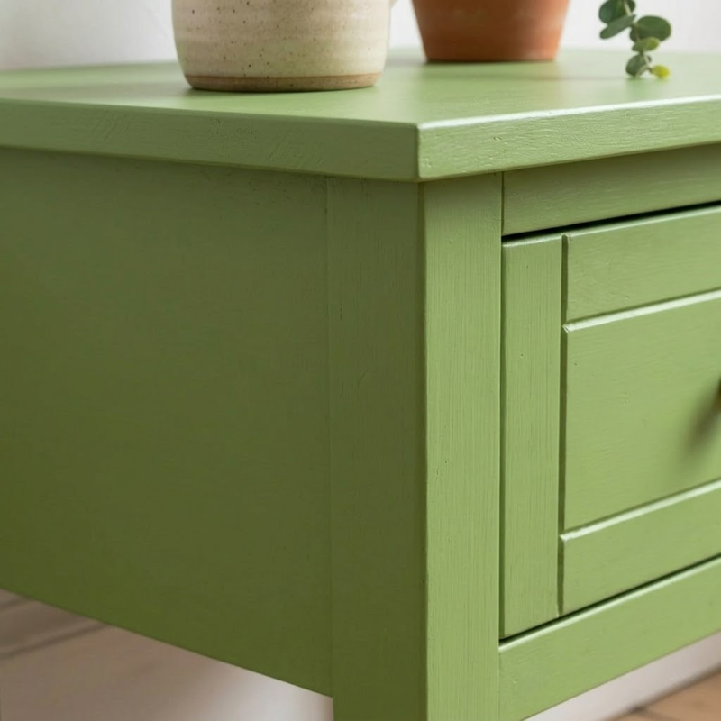 A close-up of a sage green wooden side table or cabinet with a single visible drawer front, shown in a real interior setting with potted plants on top.