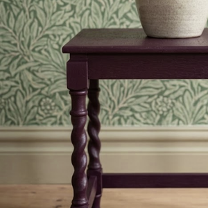 
                      
                        A close-up of a dark purple wooden table leg and tabletop corner, with a textured cream pot on top and a green botanical patterned wallpaper in the background.
                      
                    