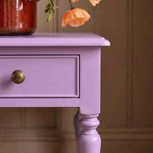 
                      
                        A close-up of a lavender purple wooden side table with a single drawer and a round brass knob, shown in an indoor setting with a vase and flowers on top.
                      
                    