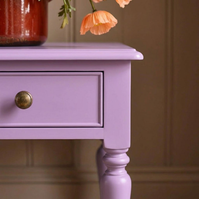 A close-up of a lavender purple wooden side table with a single drawer and a round brass knob, shown in an indoor setting with a vase and flowers on top.