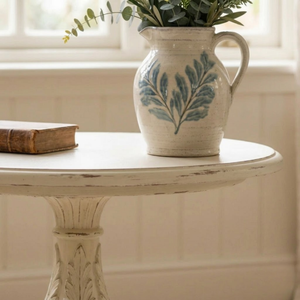 
                      
                        A distressed white ceramic pitcher with a blue leaf pattern, filled with green eucalyptus stems, sitting on a matching white round table next to a vintage book.
                      
                    