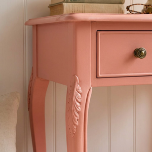 
                      
                        A close-up of a dusty pink wooden writing desk with a single drawer, ornate carved legs, and a round vintage-style metal knob, set against a white paneled wall.
                      
                    