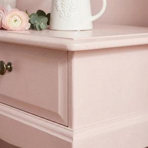
                      
                        A close-up of a dusty pink wooden nightstand with a single drawer and a dark metal knob, styled with a white mug and pink flowers on top.
                      
                    