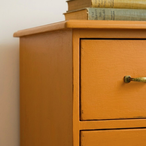 
                      
                        Close-up of a painted wooden dresser or cabinet corner with a warm orange-brown finish, a single brass-toned drawer pull, and books stacked on top against a plain light wall.
                      
                    