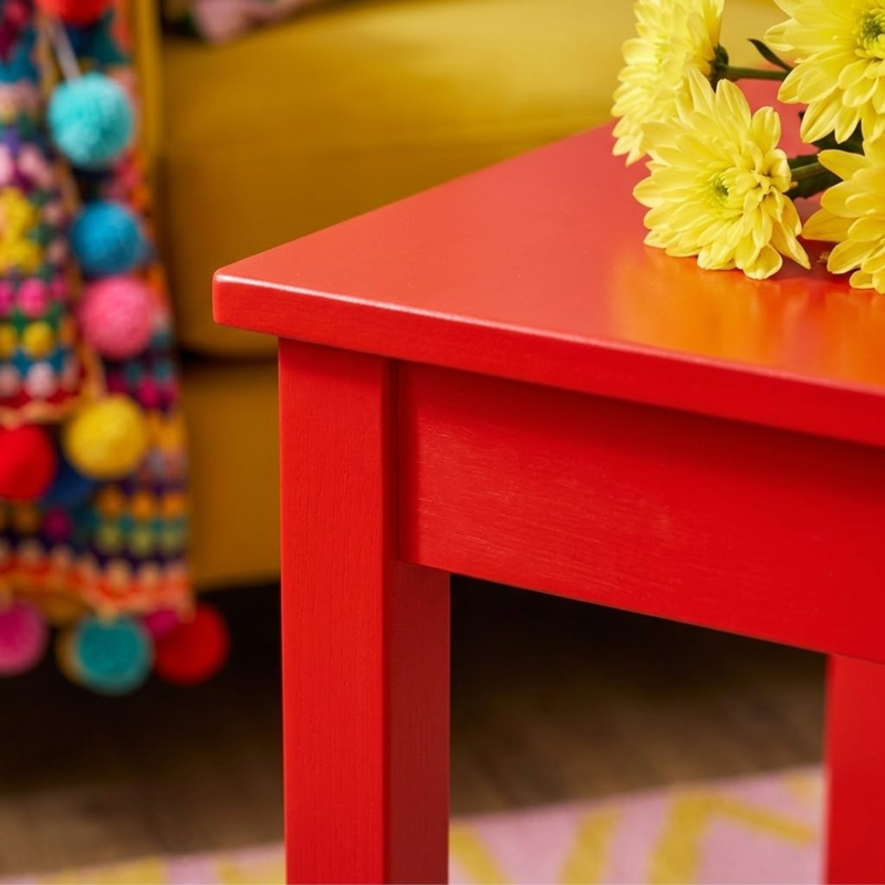 A bright red wooden side table in a cozy room setting, styled with yellow flowers on top and a colorful pom-pom bag in the background.