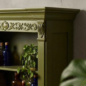 
                      
                        An olive green wooden hutch or cabinet with ornate carved detailing on the upper frame, shown in a styled interior setting with glass bottles and a trailing green plant.
                      
                    