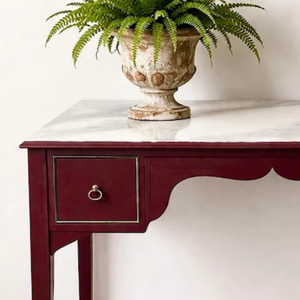 
                      
                        A dark red console table with a white marble top and a single drawer, styled with a green fern in a distressed ceramic pot against a white wall.
                      
                    
