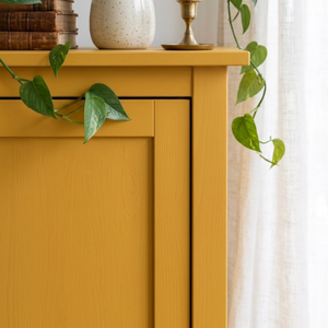 
                      
                        A mustard yellow wooden cabinet or sideboard shown in a styled interior setting, with green trailing vines draped over it, a ceramic vase, books, and a brass candlestick on top, next to a white curtain.
                      
                    