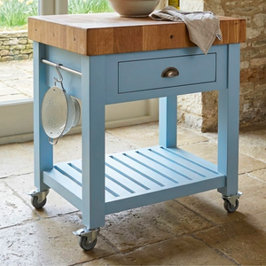 
                      
                        A light blue rolling kitchen island cart with a thick wooden butcher-block style top, a single drawer with a curved metal pull, an open lower shelf, and caster wheels. A metal colander hangs from a side hook, and a beige cloth and bowl are placed on the wooden top, set in a rustic kitchen interior.
                      
                    