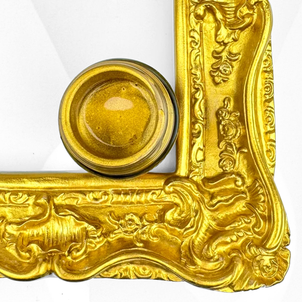 A small round glass jar filled with shimmering gold powder or pigment, placed on the corner of an ornate gold picture frame against a white background.