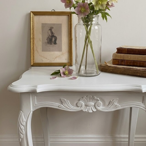 
                      
                        A white carved console table styled with a gold-framed sketch, a tall glass vase holding pale pink and green flowers, and a stack of vintage books against a neutral wall.
                      
                    
