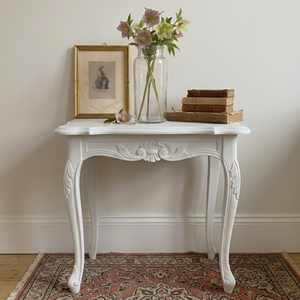
                      
                        A white carved console table with cabriole legs, styled with a gold-framed picture, a glass vase with flowers, and a stack of vintage books, set against a neutral wall on a patterned rug.
                      
                    