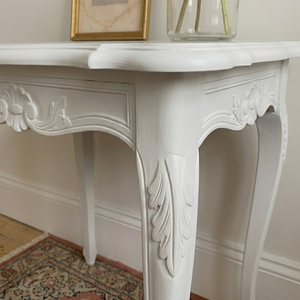 
                      
                        A white carved console table with ornate cabriole-style legs, shown in a styled interior setting with a framed picture and a glass vase on top.
                      
                    