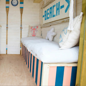 
                      
                        A nautical-themed wooden daybed with striped pink, blue, and white sides, white cushions, and decorative pillows including a lobster and starfish design, set against a light wood-paneled wall with oars and a 'BEACH' sign.
                      
                    