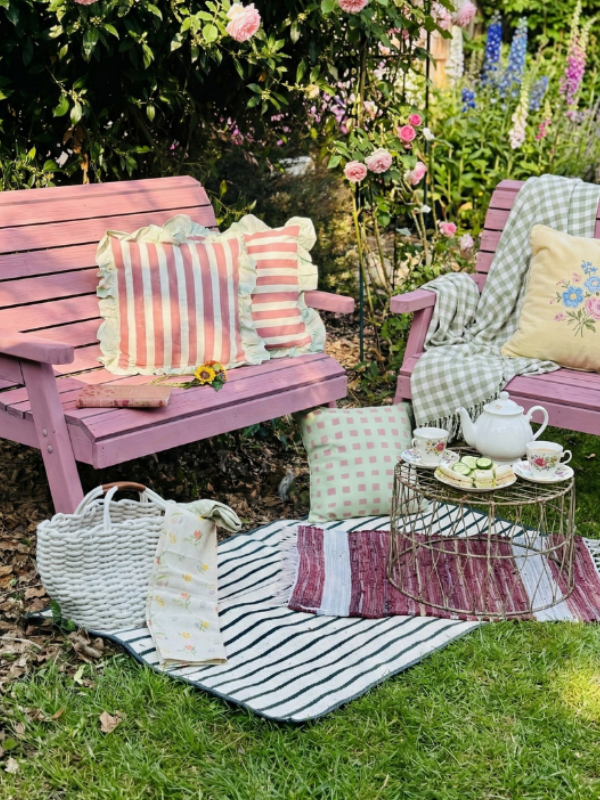 Pink garden bench with cushions and a teacup set on a striped blanket in a garden setting.