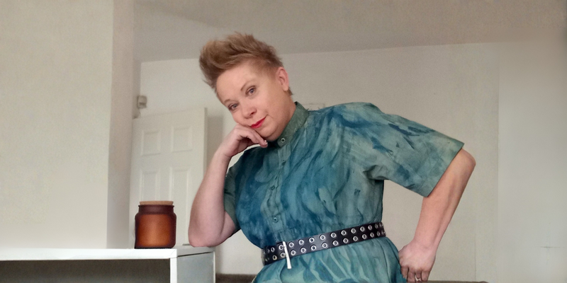 Woman in a teal dress posing in a room with a white wall and a brown jar on a shelf.