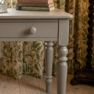 
                      
                        A close-up of a light gray painted wooden side table with a single drawer and turned legs, shown in a styled room setting with patterned curtains, books, and a wooden floor.
                      
                    
