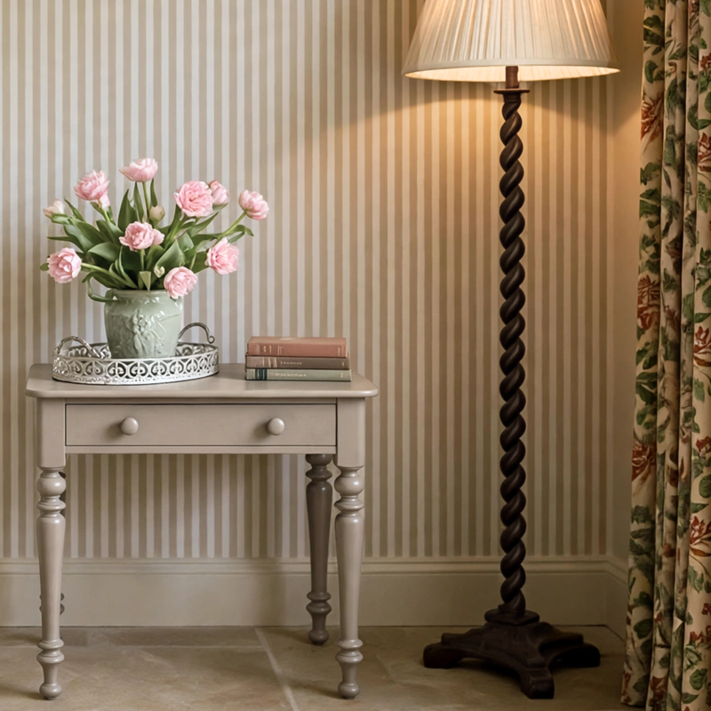 A styled interior scene showing a tall dark twisted-wood floor lamp beside a pale gray single-drawer table holding pink tulips and books, against striped wallpaper and a floral curtain.