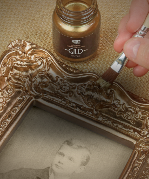 Person using a brush to apply gold paint to an ornate frame, with a bottle of gold paint labeled 'Gild' in the background.