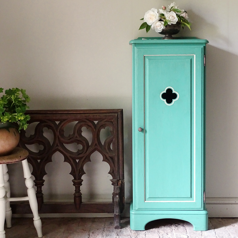 Verdigris cabinet with decorative door in a room with a wooden bench and plants.