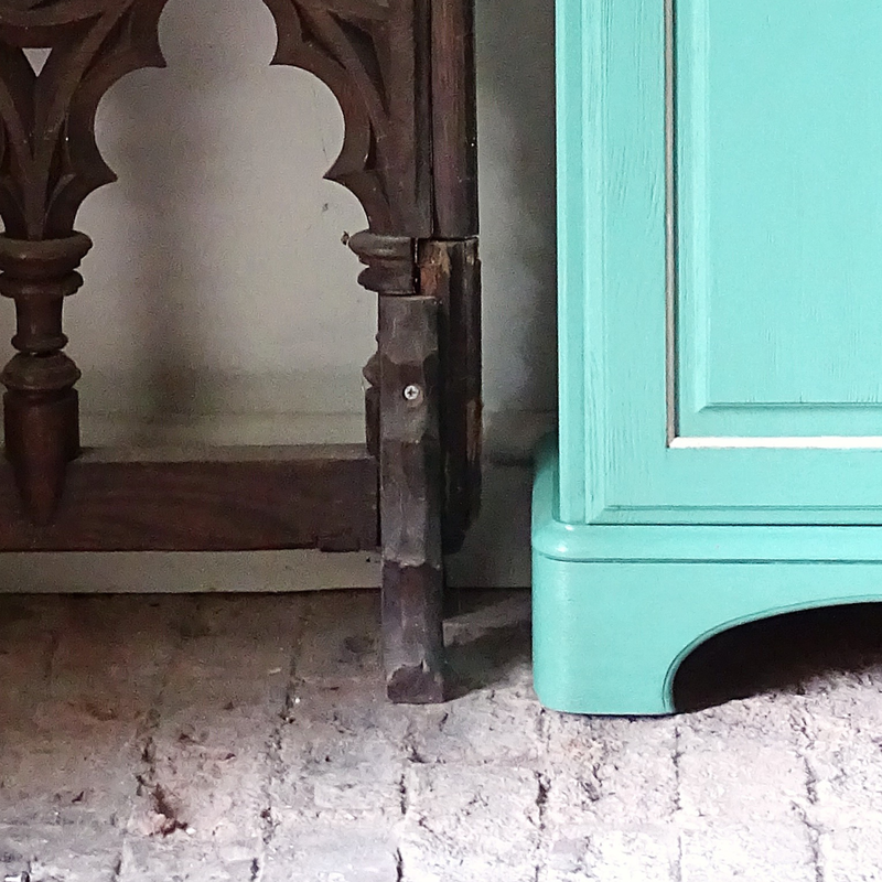 Close-up of a wooden leg and a Verdigris cabinet on a textured floor.