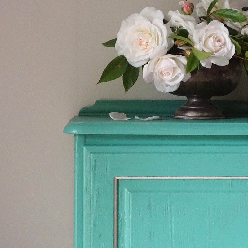 Verdigris cabinet with a vase of white flowers on top against a plain background