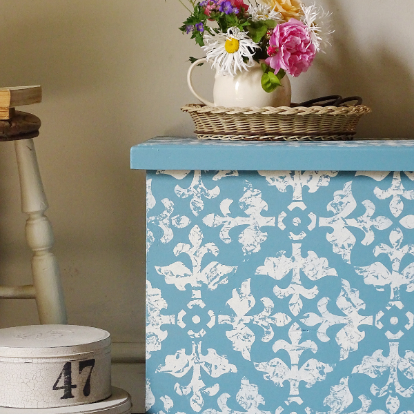 A decorative storage chest with a light blue and white distressed damask pattern, styled with a wicker basket and a white pitcher of flowers on top.