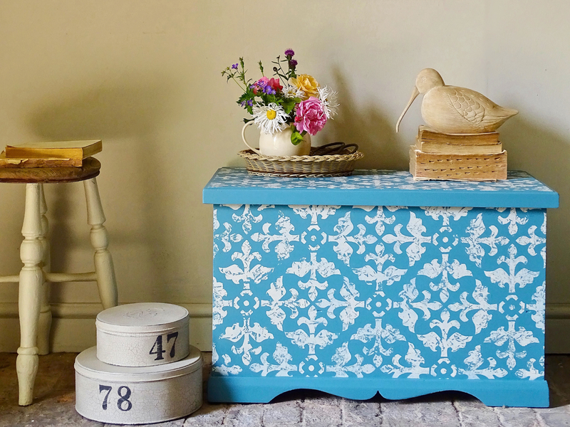 A wooden blanket box painted blue with a white stencilled pattern, beside a cream stool and stacked hat boxes