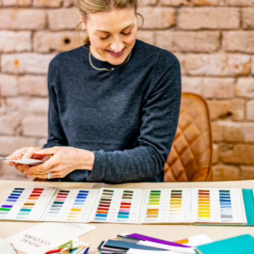 A woman in a navy blue sweater sits at a table looking through color swatch cards in front of a brick wall.
