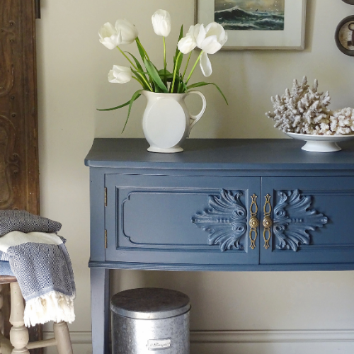 A blue console table styled in a home interior with a white pitcher of tulips, a bowl of coral, and a gray throw blanket on a chair.