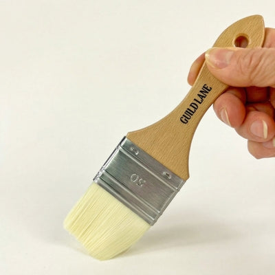 Paintbrush with wooden handle and visible brand name 'GUILD LANE' held by a hand against a plain background.