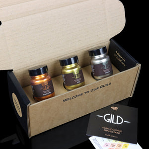 
                      
                        Three jars of metallic paint in gold, burnt copper, and platinum colors, placed inside an open box with a welcome message. A card outside the box indicates the product line 'GILD'.
                      
                    