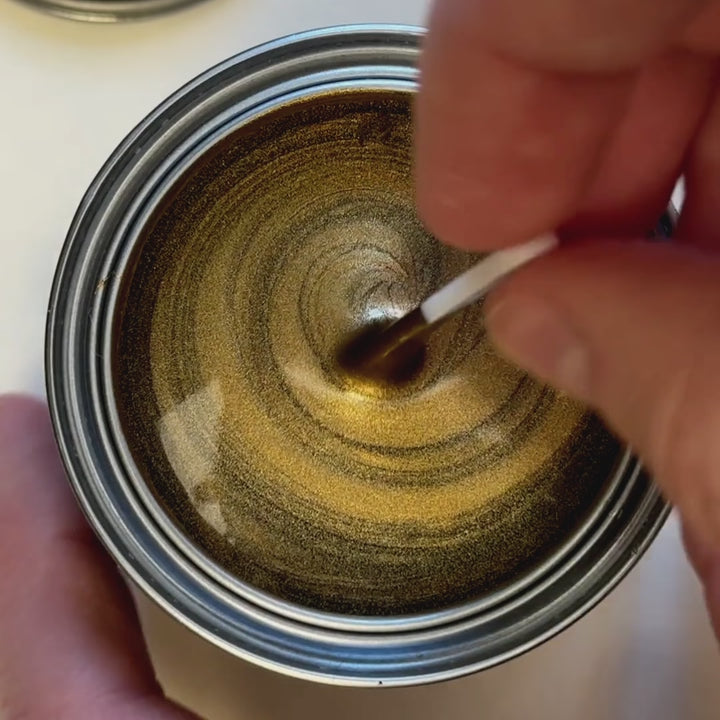 A close-up video showing a hand stirring a rich, deep bronze metallic paint in a small tin. The paint is then swatched onto a white surface with a brush, displaying a smooth, reflective finish next to an ornate gold frame.