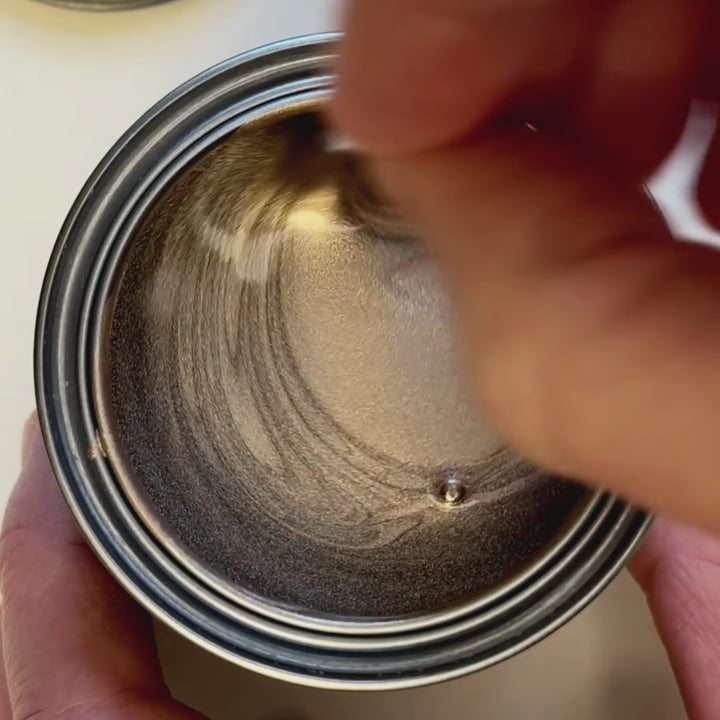 A hand stirs a pale, sophisticated champagne-coloured metallic paint, creating swirling patterns in the tin. The brush swatches the liquid onto paper, revealing a soft, pearlescent glow with high-shine metallic pigment.