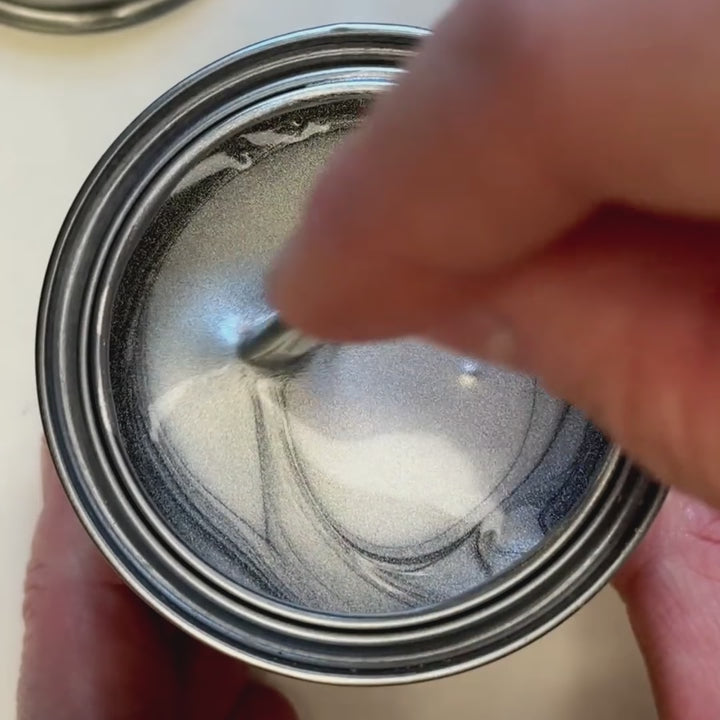 A hand stirs a bright, liquid silver metallic paint. The video captures the paint being swiped across a surface next to a silver-leafed ornate frame, showcasing a brilliant, mirror-like metallic finish.