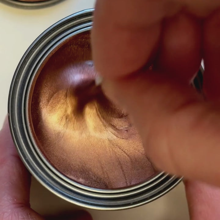 A video of rose gold metallic paint being mixed, showing a blend of pink and gold pigments. A paintbrush applies a smooth swatch to a surface, demonstrating its high-pigment coverage and metallic sheen.